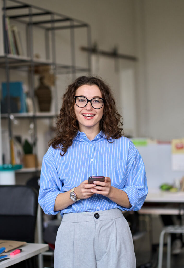 Happy young business woman holding phone standing in office, vertical.
