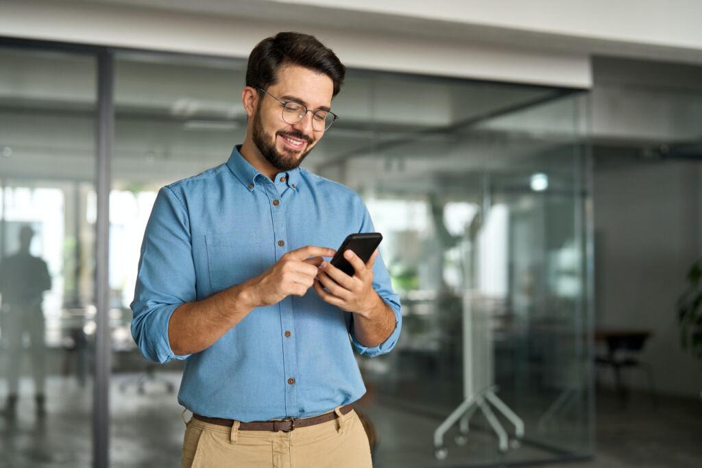 Happy smiling young latin business man holding smartphone standing in office.